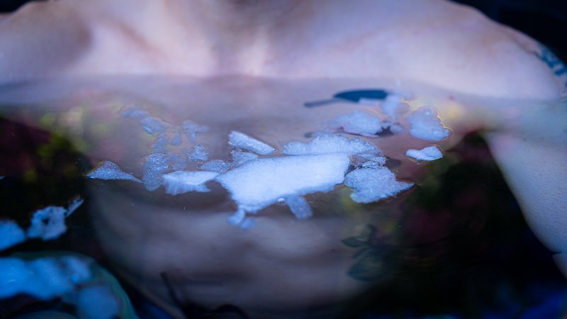 Torace nel bagno di ghiaccio con pezzi di ghiaccio sulla superficie dell'acqua Primo piano del torace di una persona durante un bagno di ghiaccio, pezzi di ghiaccio galleggiano sulla superficie dell'acqua davanti al petto.