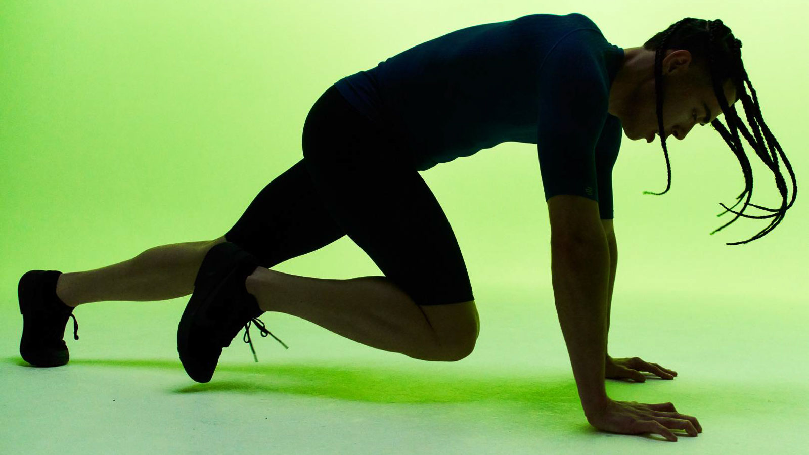Un modello fitness maschile con pantaloncini a compressione neri e una maglia abbinata, che esegue mountain climbers contro uno sfondo verde vivace.
