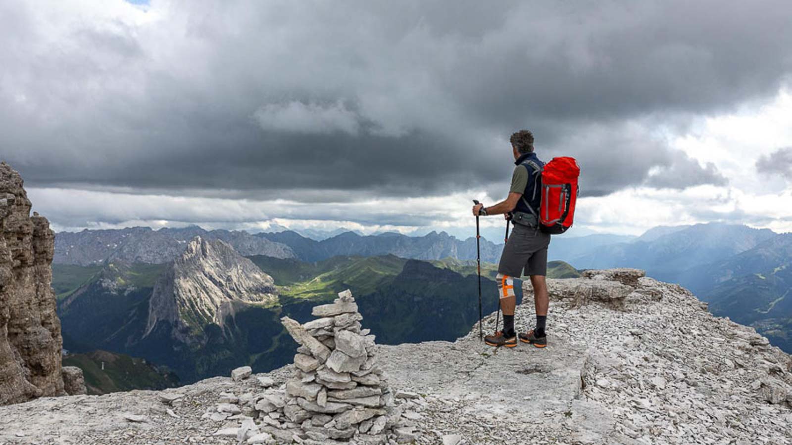 Escursionisti su un promontorio roccioso con una ginocchiera da trekking Una persona con uno zaino rosso è in piedi su un affioramento roccioso nelle Dolomiti, guardando lontano. Il cielo è nuvoloso e c'è una costruzione di ometti di pietra in primo piano.