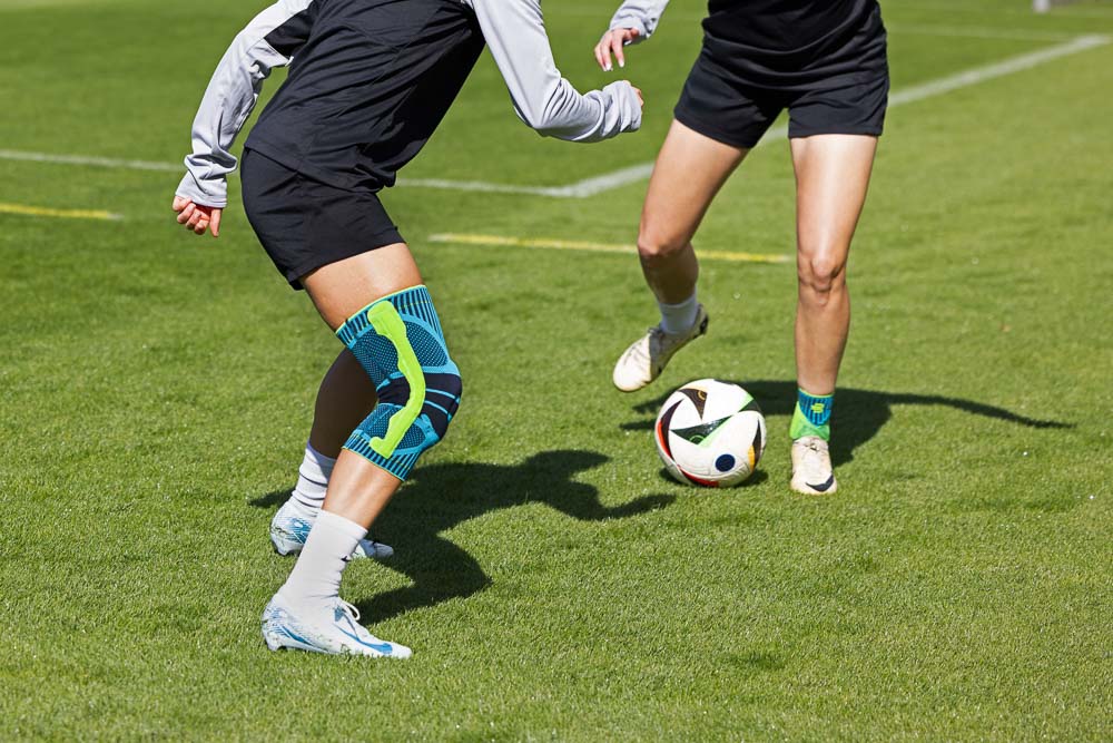 Due calciatrici si sfidano. Una indossa una ginocchiera Bauerfeind Sports, l'altra una cavigliera Bauerfeind Sports.