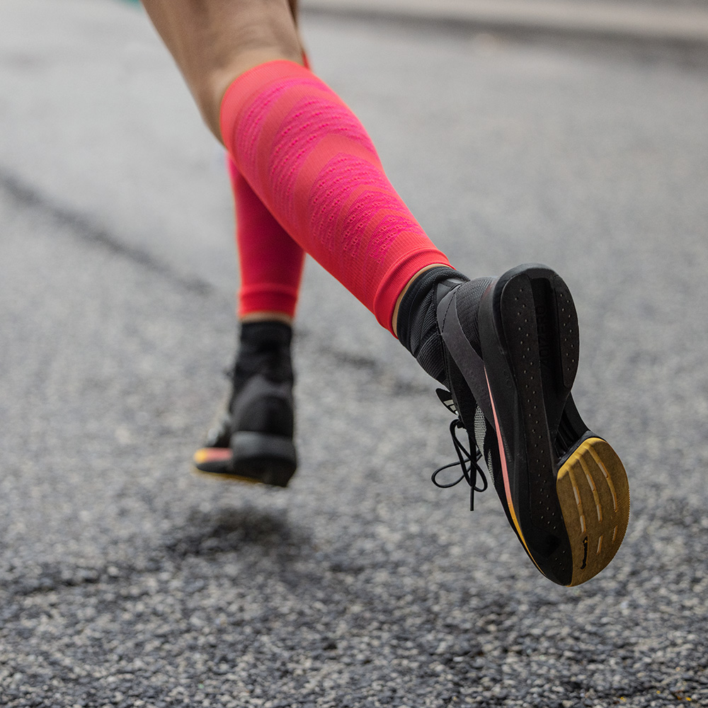 Dettaglio di una runner in movimento su asfalto, ripresa da dietro. Indossa scarpe da corsa nere e vistose Run Performance Sleeves rosse con compressione per sostenere i muscoli del polpaccio.