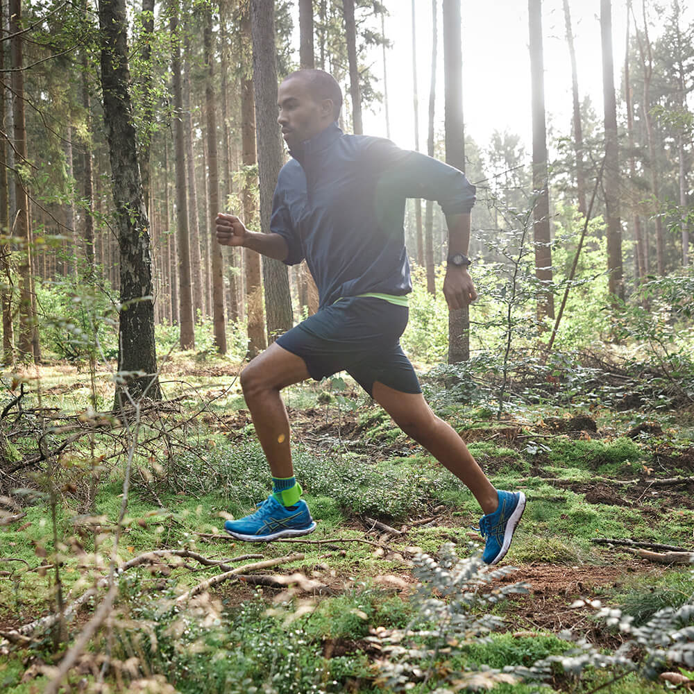 Un uomo corre dinamicamente attraverso una foresta soleggiata. Indossa una giacca blu, pantaloncini neri e fasce per i polpacci verde brillante.