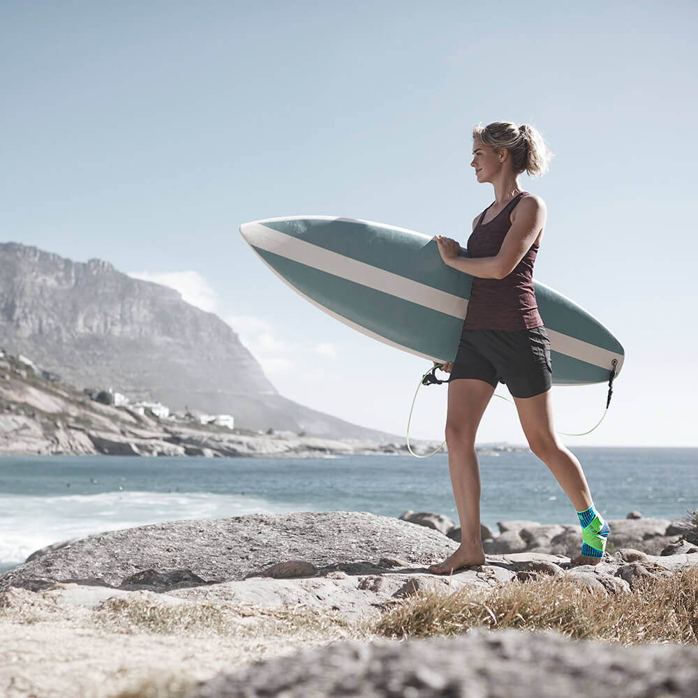 Donna con una tavola da surf sotto il braccio con una benda alla caviglia sulla gamba sinistra su una spiaggia pietrosa con mare e montagne sullo sfondo