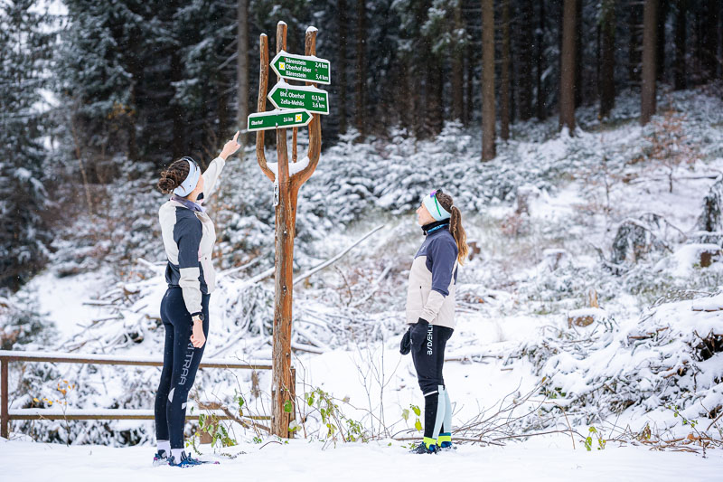 Trovare il percorso giusto Due donne in abbigliamento da corsa invernale si trovano davanti a un cartello di legno nella foresta innevata. Una indica le frecce mentre l'altra osserva attentamente. Il cartello mostra vari percorsi escursionistici.