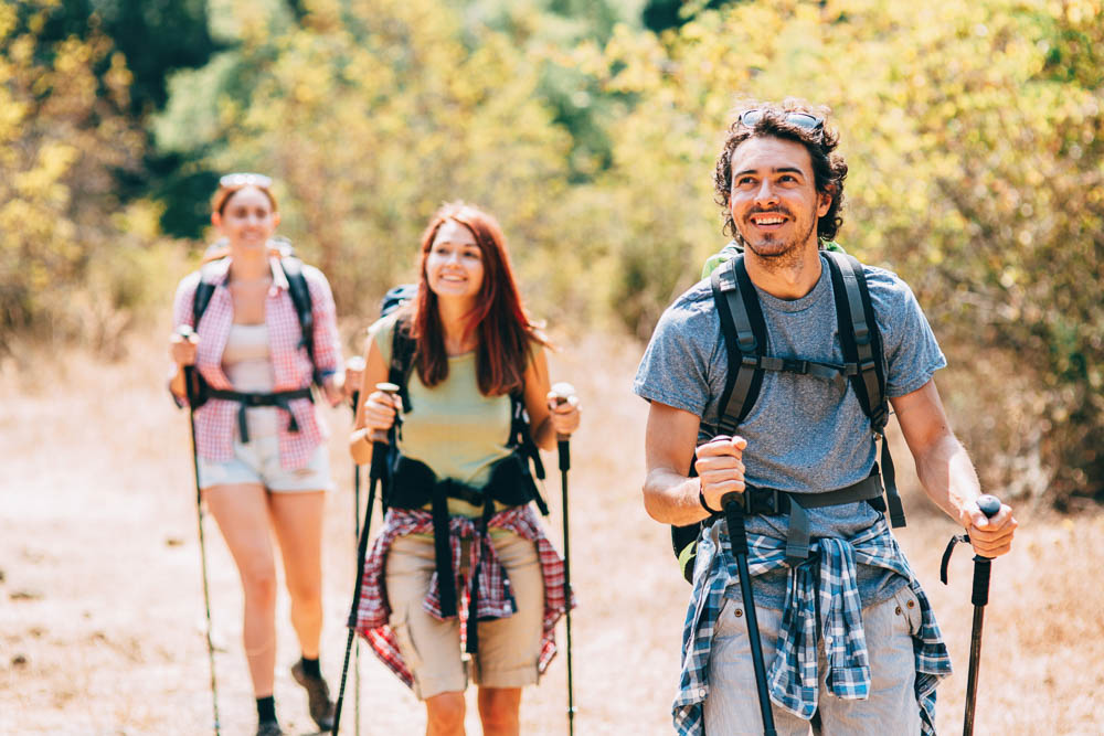 Piacere dell’escursionismo su sentiero soleggiato Tre escursionisti, due donne e un uomo, camminano felici con bastoncini da trekking su un sentiero boschivo soleggiato. Indossano abbigliamento sportivo e zaini – il focus è sul movimento naturale e sull’esperienza all’aperto condivisa.