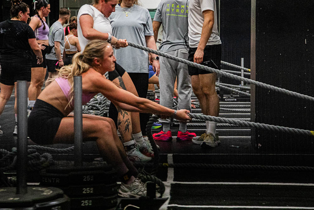 Atleta impegnata nello sled pull durante una gara HYROX, mentre tira con una lunga corda una slitta pesante sul pavimento indoor; sullo sfondo si vedono staff e altri partecipanti vicino alle stazioni di workout, in una scena che esprime forza,