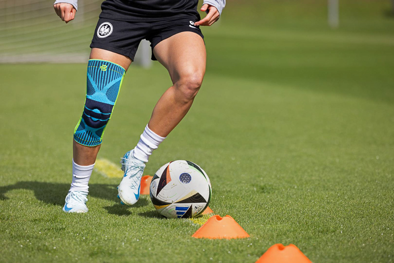 Una calciatrice si allena sul campo indossando un tutore per ginocchio Bauerfeind. L'attenzione è sulla stabilità e sul supporto forniti dal tutore durante i movimenti dinamici, come il dribbling intorno ai coni.