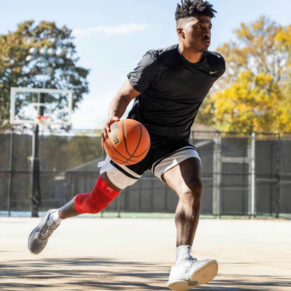 Ein Basketballspieler mit roter Kniebandage sprintet über den Außenplatz, während er den Ball kontrolliert. Der sonnige Tag und die Bäume im Hintergrund verleihen der Szene eine dynamische Atmosphäre.