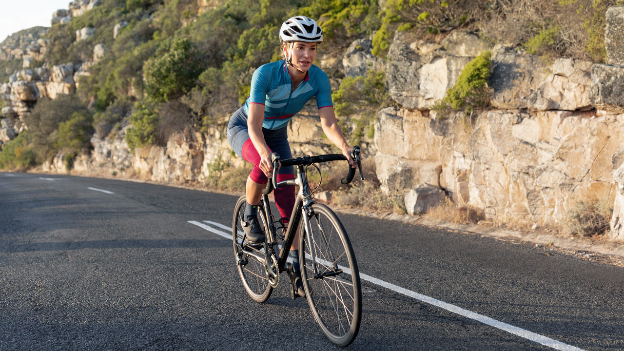 Ciclista con maglia blu e pantaloncini rosa che indossa il ginocchiera NBA Compression Knee Support, mentre pedala su una bici da corsa lungo una strada costiera con scogliere rocciose in una giornata di sole.