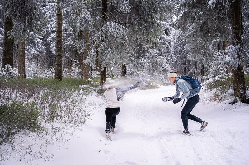 Battaglia a palle di neve – La gioia infantile nella neve fredda Due donne con abbigliamento invernale caldo si divertono con una battaglia a palle di neve in una foresta ricoperta di neve. La donna a destra indossa una giacca grigio-blu e uno zaino mentre sta per lanciare una palla di neve.