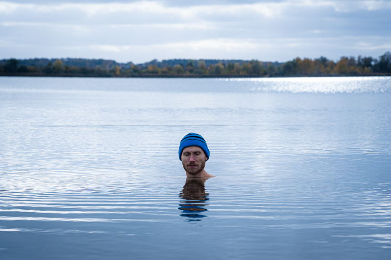Uomo nel lago durante un bagno di ghiaccio con berretto blu Un uomo si trova in un lago tranquillo, immerso fino al collo, indossa un berretto blu, e il paesaggio autunnale si riflette nell'acqua.