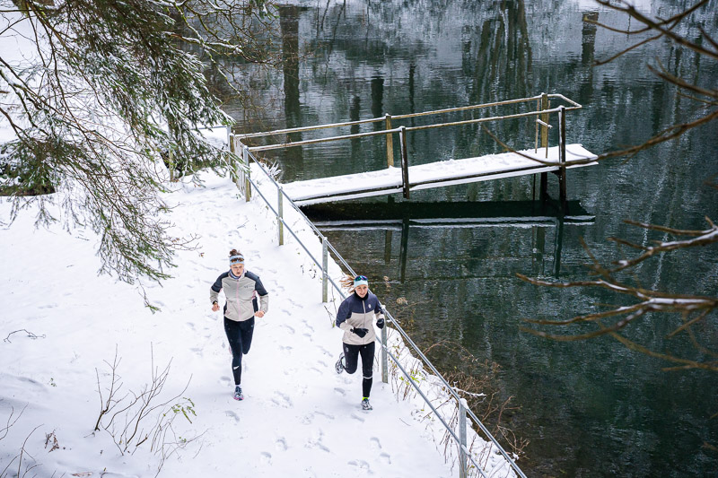 Corsa invernale al lago: Impronte nella neve, riflessi nell’acqua. Due runner corrono lungo un sentiero innevato accanto a un lago con un pontile coperto di neve. La superficie del lago riflette gli alberi circostanti. Entrambe le runner indossano giacche da corsa invernali chiare e pantaloni neri.