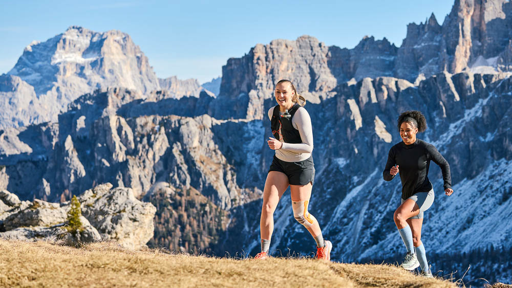 Due runner si allenano insieme su una cresta di montagna con vista su un paesaggio alpino roccioso. Una delle atlete indossa una ginocchiera compressiva outdoor. L’immagine esprime dinamismo e allenamento all’aria aperta.
