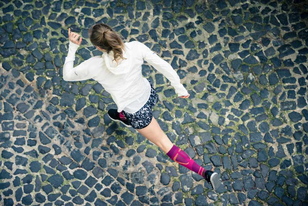 Bird's Eye View: Runner in una giacca da corsa bianca e con i calzini in corsa rosa corre su una strada della Città Vecchia