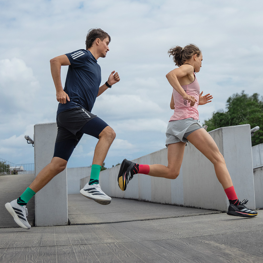 Due runner in movimento su una rampa, un uomo con pantaloncini blu e calze da running verdi Bauerfeind e una donna con pantaloncini grigi e calze rosse Bauerfeind.