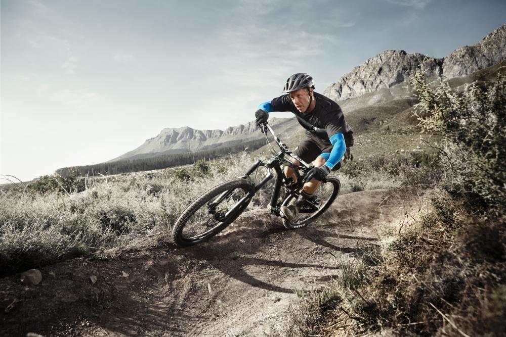 L'uomo con armi si guida su una mountain bike in un angolo estremo su un sentiero polveroso attraverso un paesaggio sterile con cespugli e montagne sullo sfondo