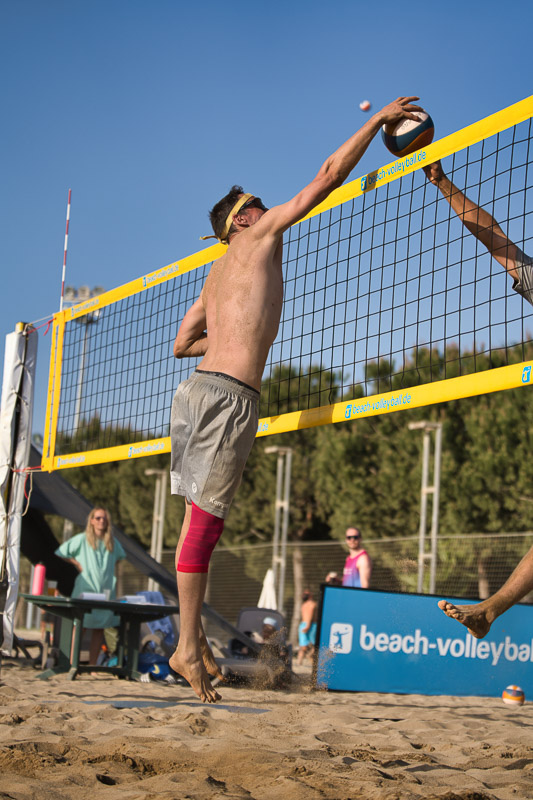 Un giocatore di beach volley salta a rete per bloccare, mentre un avversario colpisce la palla. L’atleta indossa una fascia compressiva rosso acceso sulla gamba destra.