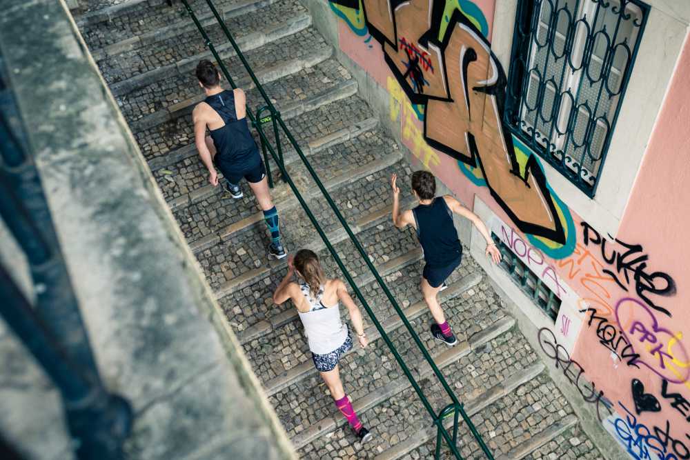 Bird's Eye View: un uomo e due donne in abiti da corsa corrono una scala in una città vecchia con graffiti sui muri