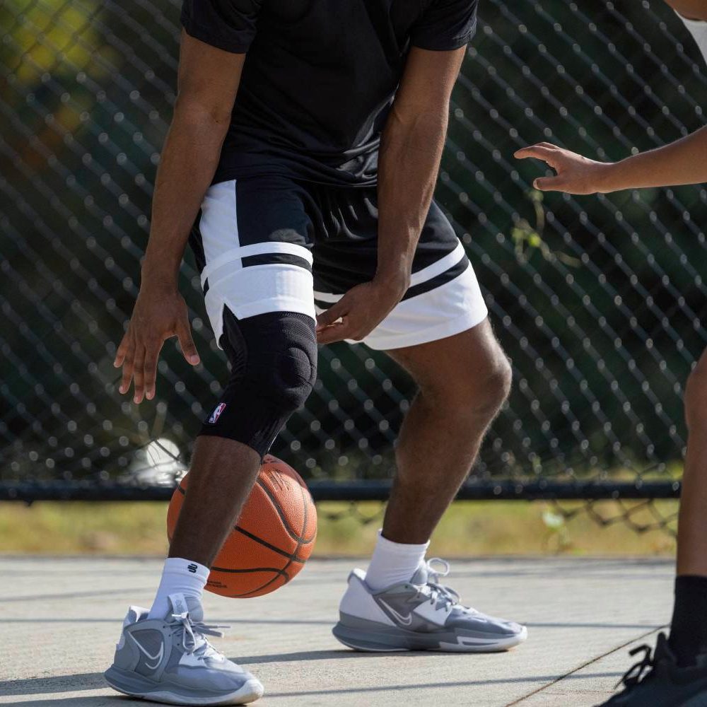 Palleggio con ginocchiera nera durante la partita Primo piano di un giocatore di basket mentre palleggia su un campo esterno. Il ginocchio destro è protetto da una ginocchiera nera con logo NBA. Un avversario è parzialmente visibile.