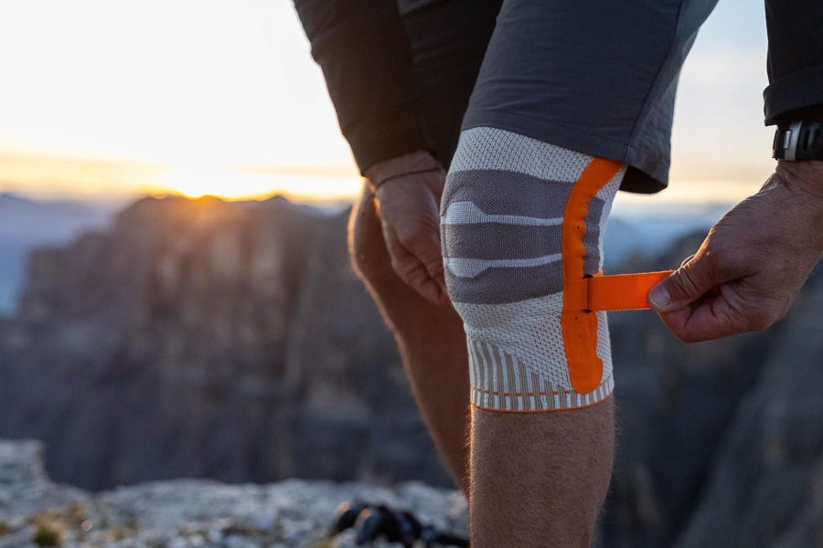 Un escursionista regola la sua ginocchiera arancione durante un'escursione in montagna. Sullo sfondo si vedono montagne rocciose e un tramonto.