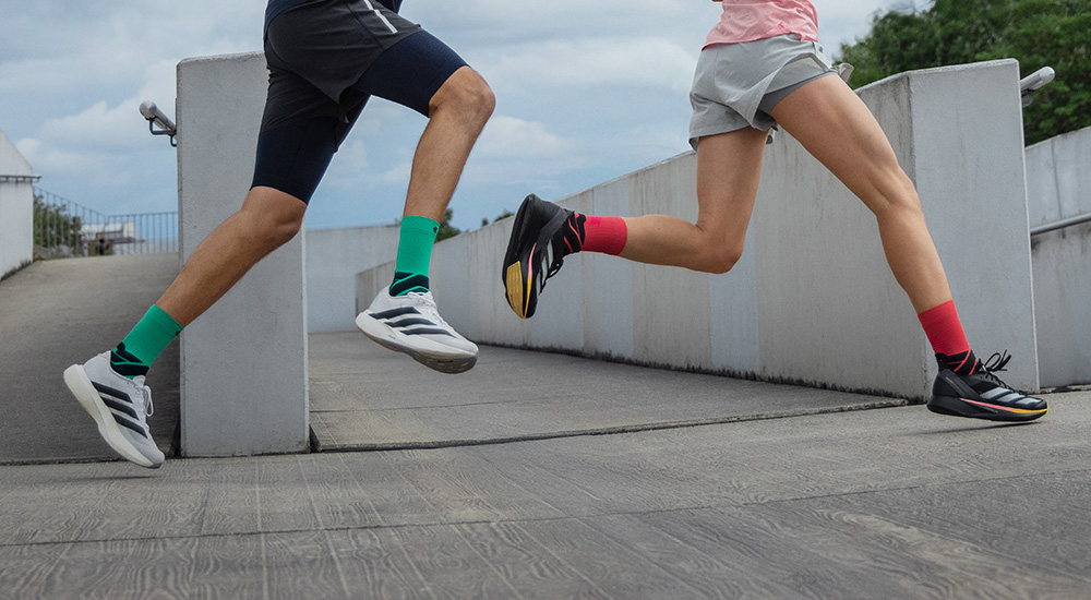 Energia in avanti Due runner in movimento che scattano su una rampa. L’uomo indossa calze a compressione verdi con scarpe da corsa bianche, la donna scarpe nere con calze a compressione rosse. Entrambi colti nel pieno dell’azione.