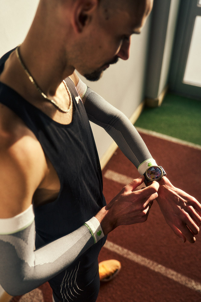 Hendrik Pfeiffer controlla il suo orologio da corsa durante l’allenamento in pista indoor, indossando maniche compressive per stabilità e performance.