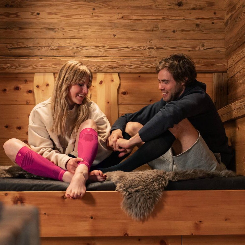 Una coppia sorridente è seduta comodamente in una baita di legno su una panca con una copertura in pelliccia. La donna indossa calze a compressione rosa, mentre l'uomo indossa quelle nere. Entrambi si godono il meritato riposo dopo una giornata attiva.