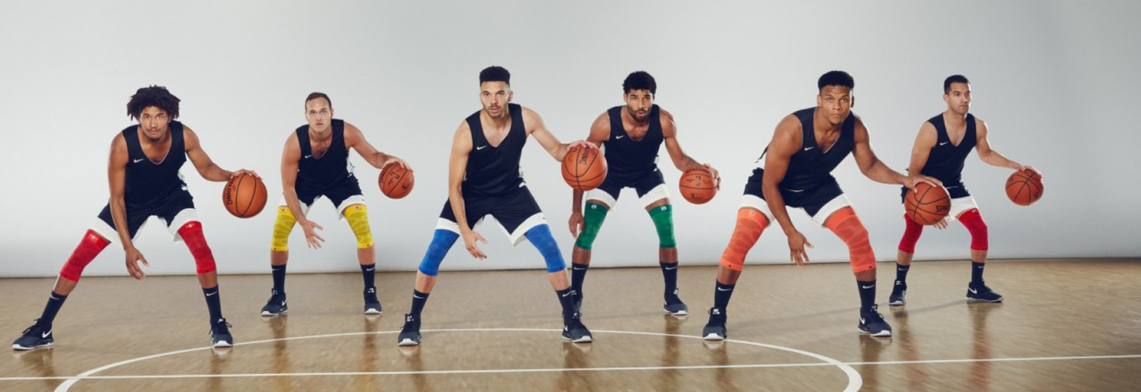 Uniti nella forza – energia e colore in campo Sei giocatori di basket sono schierati in una palestra, pronti per un'esercitazione. Indossano maglie nere, pantaloncini bianchi e fasce compressive colorate alle ginocchia. Ognuno ha un pallone da basket in mano, pronti per iniziare l’allenamento.