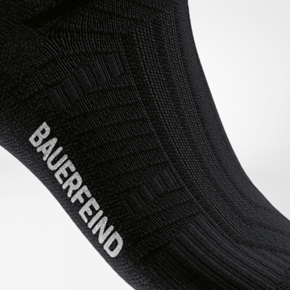 Vista laterale di una calza sportiva nera con struttura a coste nella zona mediale e logo bianco “BAUERFEIND” – per sostenere l’arco plantare durante la corsa.
