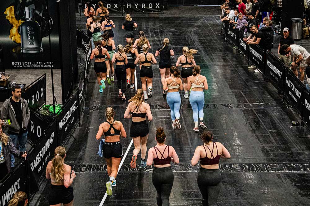 Vista dall’alto di una corsia di corsa HYROX all’interno di un’arena indoor, con numerosi atleti in abbigliamento sportivo lungo il percorso; il pubblico osserva dietro le transenne mentre i partecipanti continuano a correre con concentrazione