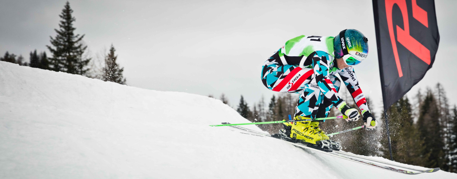 Ski Runner in una tuta da corsa blu e bianca e nero spinge una cresta ripida in accovacciarsi con le braccia davanti al corpo sullo sfondo e una bandiera di sponsor nera