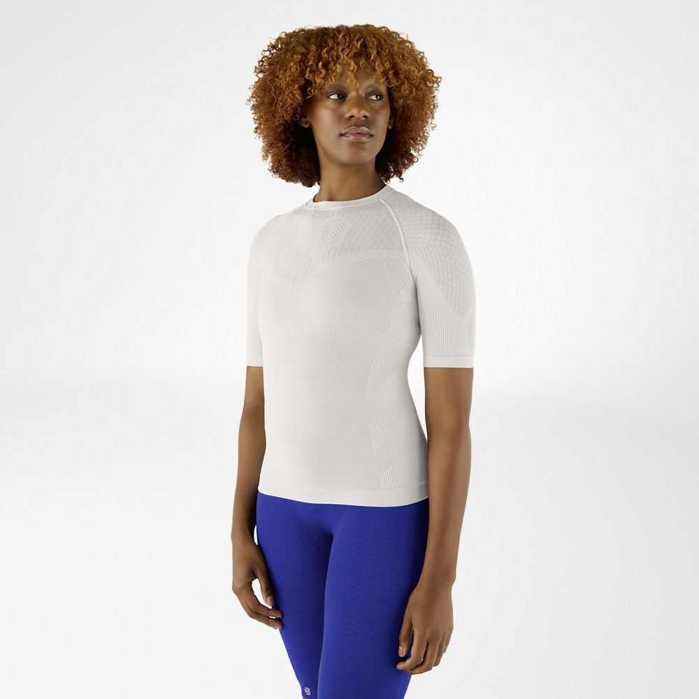 Donna con capelli ricci indossa una maglia a compressione grigio argento e leggings blu. È in piedi rivolta in avanti e guarda di lato.