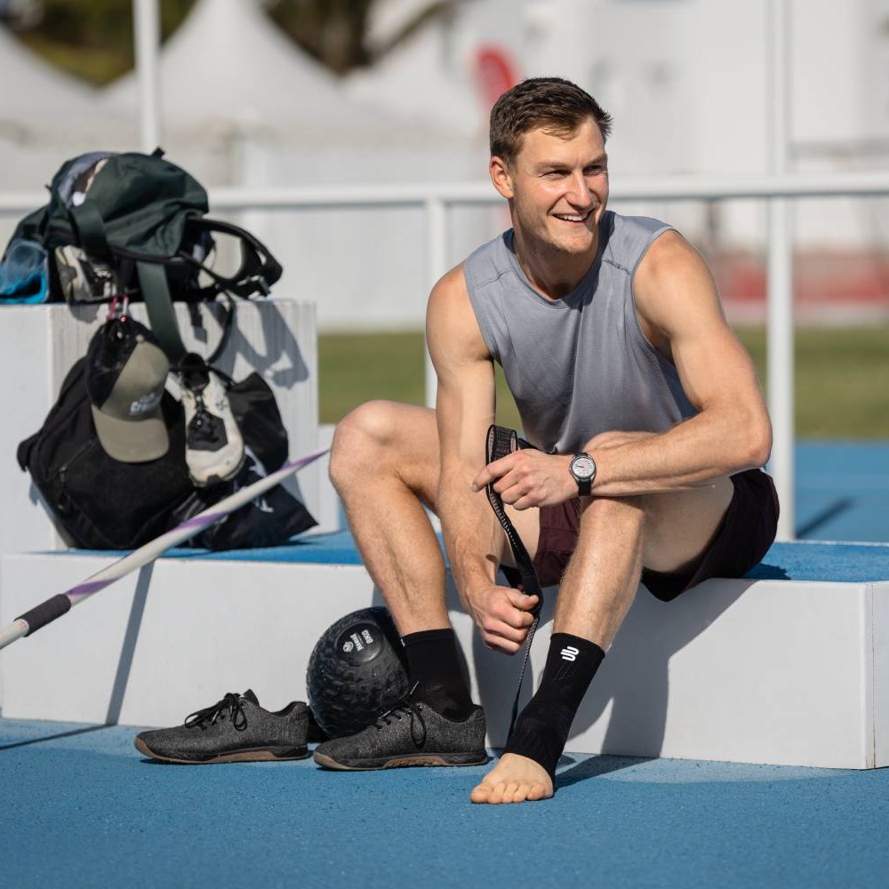 Un atleta è seduto su un podio in un impianto di atletica leggera e infila una Bauerfeind Sports Ankle Support sulla caviglia. Accanto a lui ci sono le scarpe sportive e una palla da allenamento.