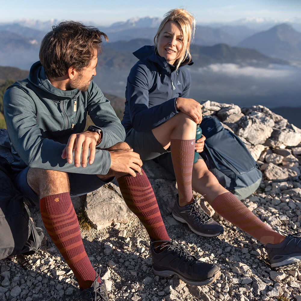 Due escursionisti godono del panorama sulla cima di una montagna, indossando calze a compressione Merino Outdoor in rosso-marrone. Le calze offrono comfort e supporto ottimali per lunghe escursioni in terreni impegnativi.