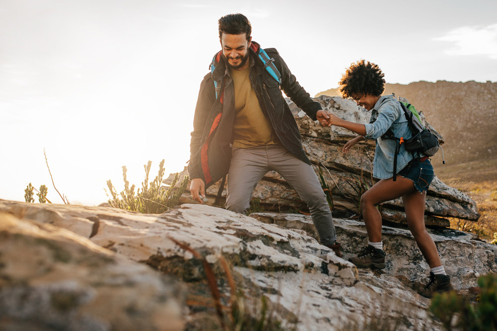 Insieme più forti sul sentiero Una giovane coppia scala insieme un sentiero roccioso al tramonto. L’uomo porge la mano alla donna per aiutarla. Entrambi indossano zaini e abbigliamento da trekking.