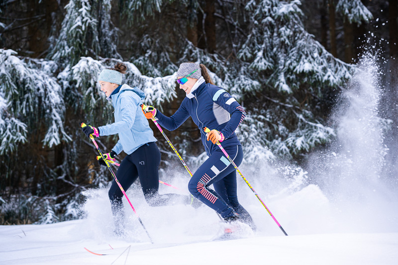 Sci di fondo dinamico nella foresta innevata Due donne che praticano sci di fondo attraverso foreste innevate. Indossano abbigliamento sportivo invernale dai colori vivaci e scivolano dinamicamente sulla neve. La scena è dominata da fitte conifere innevate, sottolineando il paesaggio invernale.