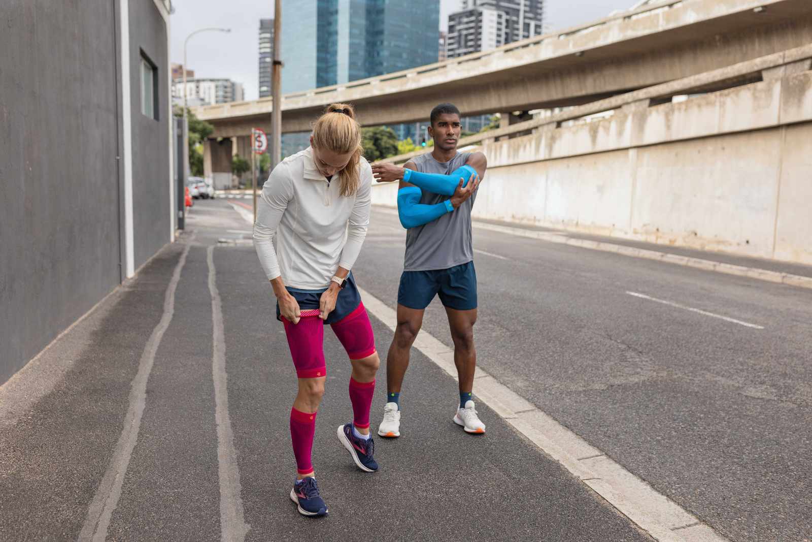 Guarda anche bene per la pausa: nessun problema con i nostri prodotti eleganti Una donna con maniche a compressione rosa e un uomo con maniche a braccio blu e calze a corto di corsa si fa una pausa sul ciglio della strada di una grande città e si allunga