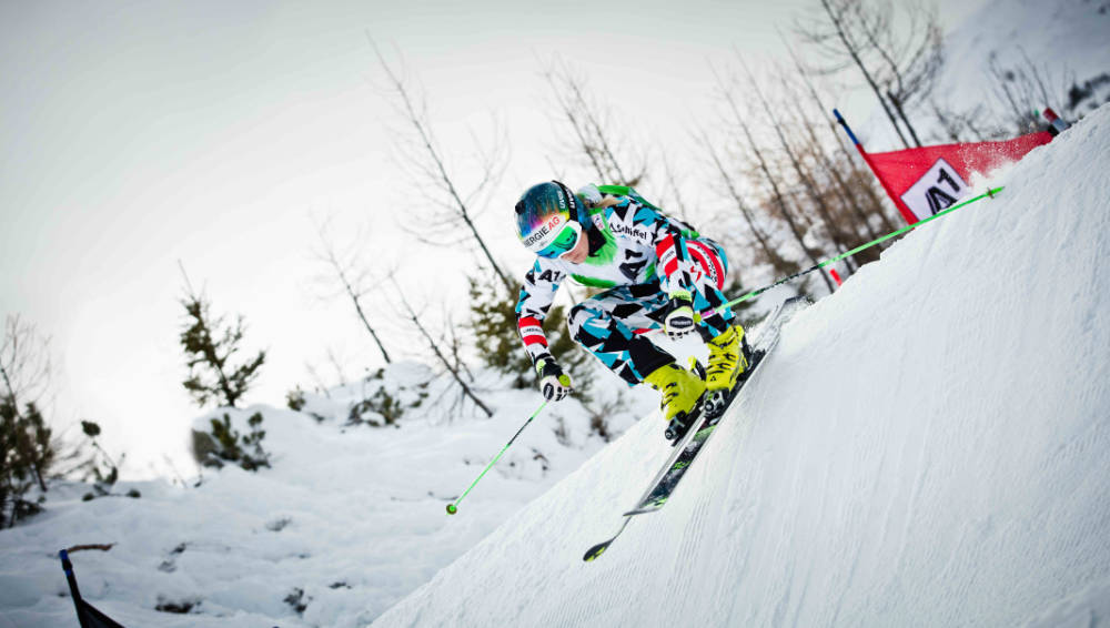 Ski Runner in una tuta da corsa blu e bianca che guida sullo sfondo un albero ripido sullo sfondo si può vedere un cancello rosso