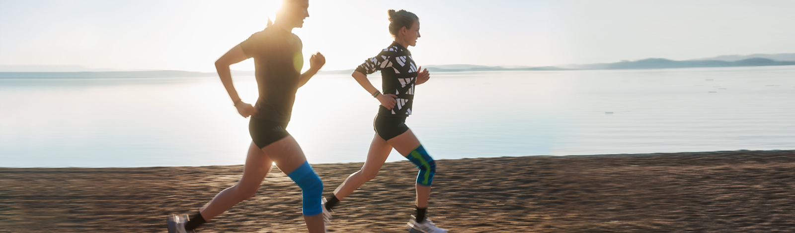 Con un ginocchio di un corridore, una fascia al ginocchio può porre rimedio alla situazione Due corridori con bande di ginocchio corrono in spiaggia accanto a un lago al Dusk