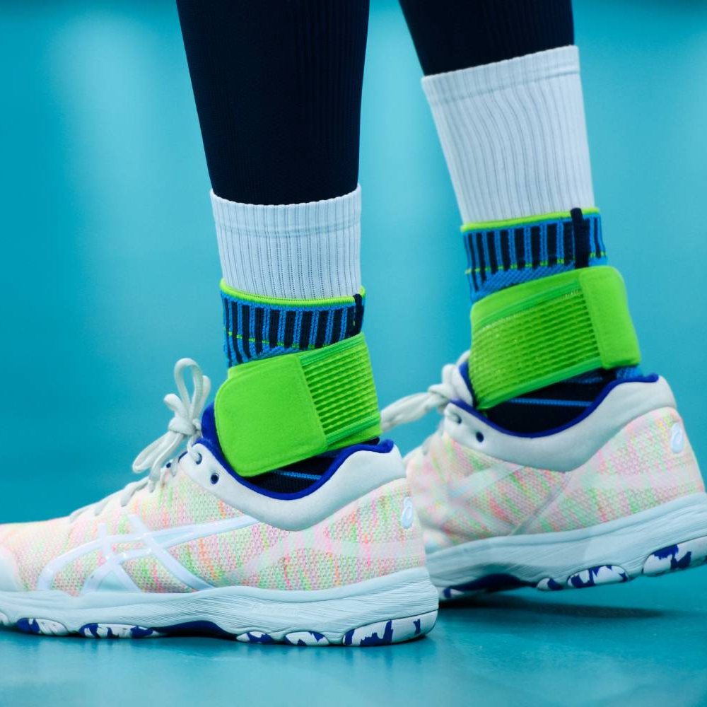 Primo piano delle gambe di un giocatore di pallavolo su un campo indoor. La Bauerfeind Sports Ankle Support Rivera è indossata sopra calzini bianchi alti.