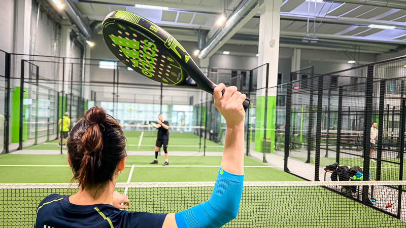 Una giocatrice di padel è posizionata a rete e si prepara a colpire la palla. Il suo braccio è protetto da una fascia compressiva azzurra.