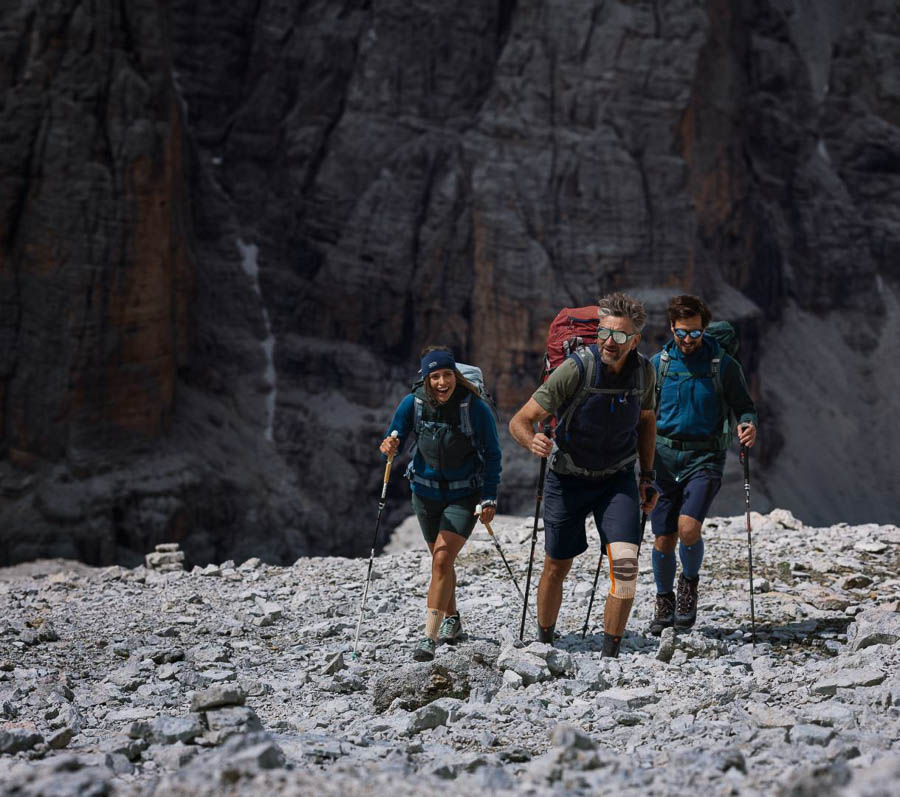 Un gruppo di tre escursionisti, equipaggiati con zaini e bastoncini da trekking, attraversano un terreno roccioso in montagna. Uno degli escursionisti indossa una ginocchiera. Sullo sfondo si vedono pareti rocciose ripide.