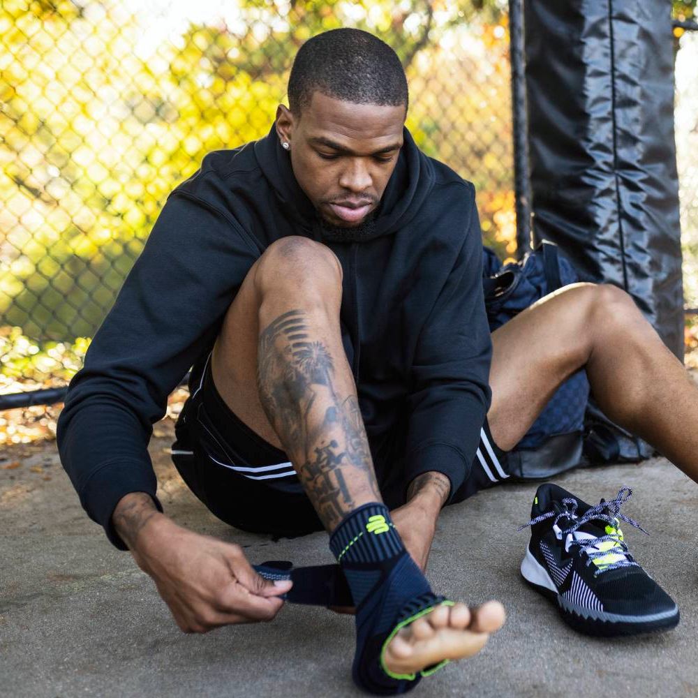 Un atleta è seduto all'aperto e avvolge una Bauerfeind Sports Ankle Support attorno alla caviglia. Le sue scarpe sportive sono accanto a lui, con una recinzione metallica sullo sfondo.