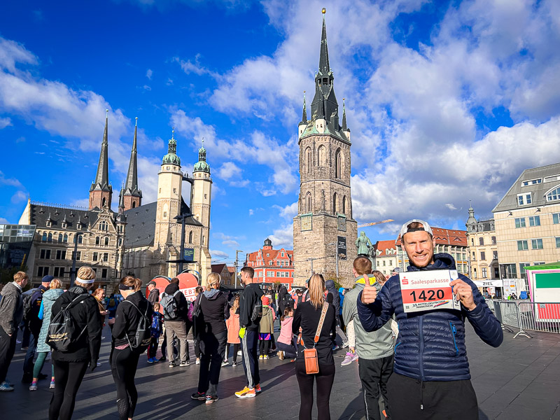 Un partecipante alla Mitteldeutscher Halbmarathon alza il suo numero di pettorale nella piazza del mercato di Halle (Saale). Dietro di lui si vedono la Torre Rossa e la Marktkirche. In primo piano, corridori e spettatori si preparano alla gara.