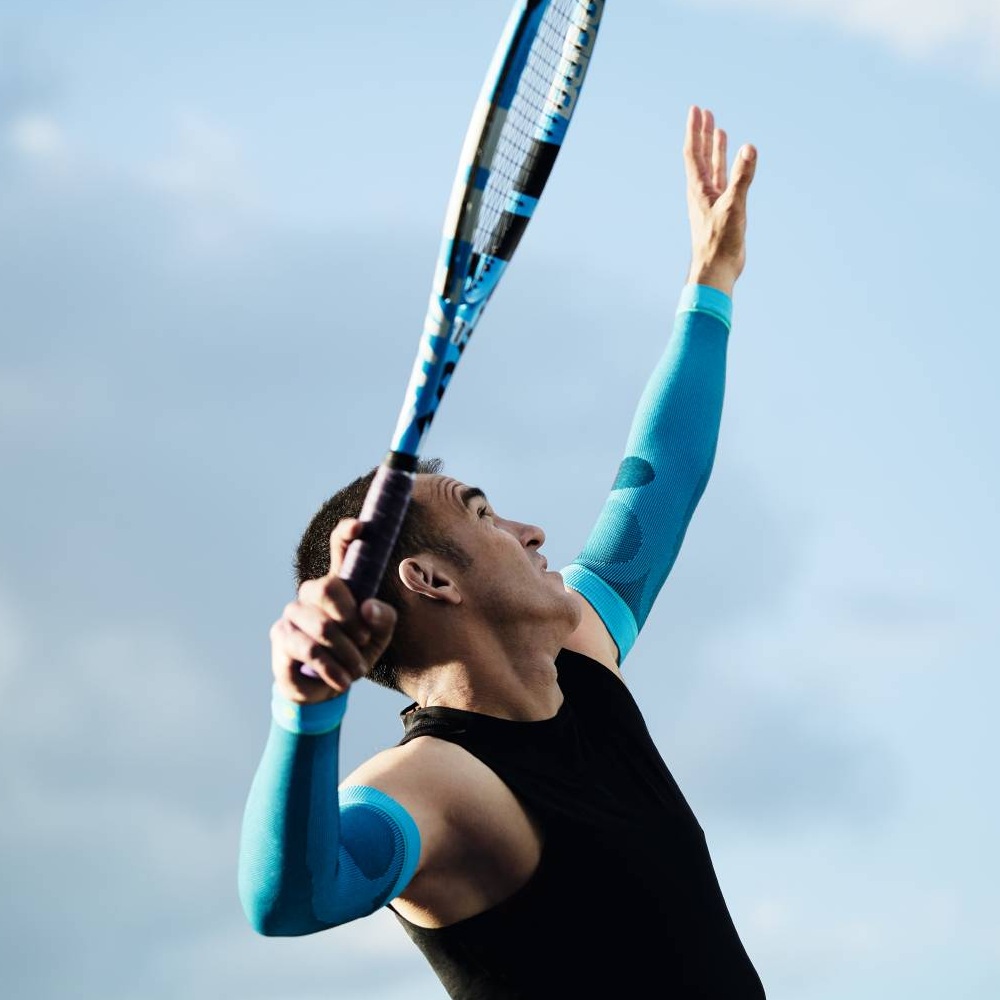 Un tennista con maniche a compressione blu sta servendo. Il cielo è azzurro con alcune nuvole.