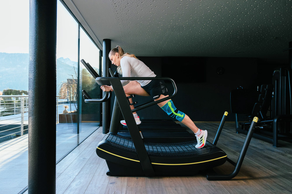 Atleta che si allena su un tapis roulant moderno in un ambiente fitness luminoso. Indossa una ginocchiera sulla gamba destra che offre stabilità e supporto durante una sessione di corsa intensa. Sullo sfondo si vede un paesaggio montano.