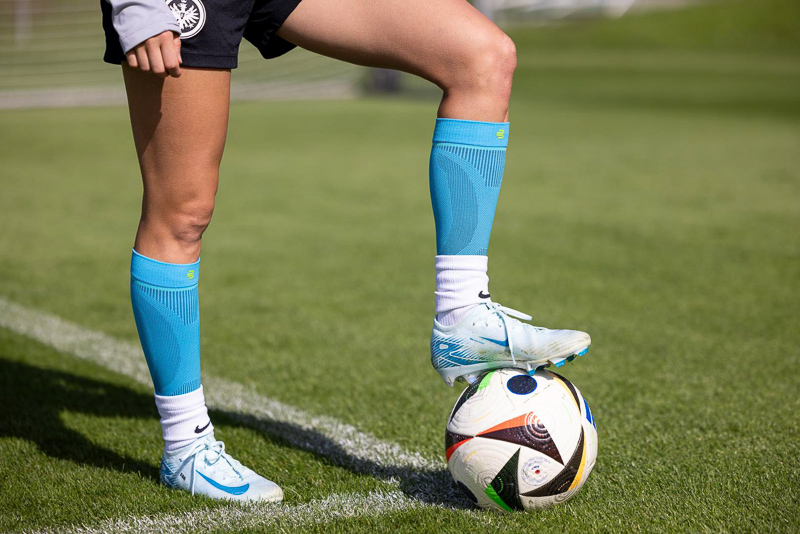 Primo piano di una calciatrice che indossa calzini a compressione Bauerfeind, con un piede appoggiato su un pallone. Il prato verde e i dettagli nitidi mettono in risalto le prestazioni atletiche e la qualità dell'attrezzatura.