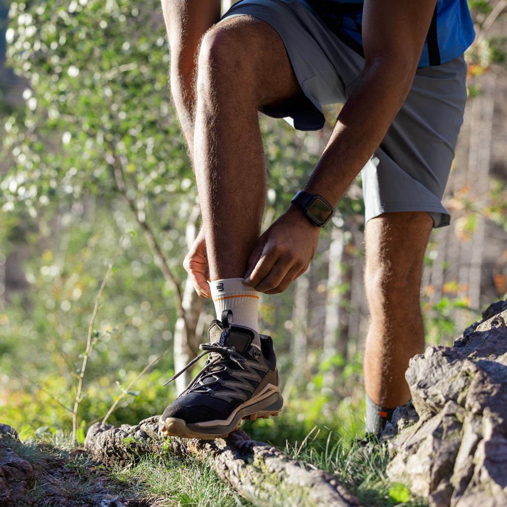 Un escursionista si trova su un sentiero nel bosco mentre sistema l’Outdoor Compression Ankle Support attorno alla caviglia. Indossa scarponi da trekking, pantaloncini e uno zaino.