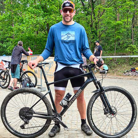 Sorriso gravel: dopo una sfida, ne arriva sempre un’altra! Un uomo sorridente posa con una bici gravel Cannondale nera su un sentiero forestale. Indossa una maglia blu a maniche lunghe Patagonia e pantaloncini da ciclismo neri. Altri ciclisti e alberi verdi sono visibili sullo sfondo.
