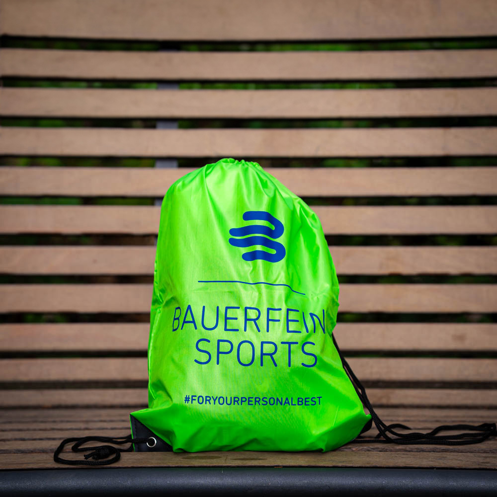 Pratico e stiloso: Lo zaino Bauerfeind Sports per i tuoi obiettivi Uno zaino verde Bauerfeind Sports è posato su una panchina di legno, mostrando il logo e l'hashtag #foryourpersonalbest.